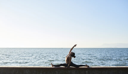 Junge Frau übt Yoga an einer Mauer am Meer - IGGF00441