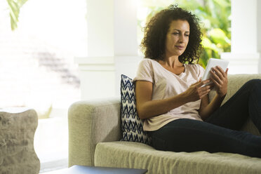 Frau mit lockigem Haar auf dem Sofa, die mit einem E-Reader liest - SBOF01444