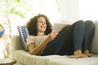 Happy woman lying on sofa with earphones and tablet - SBOF01447