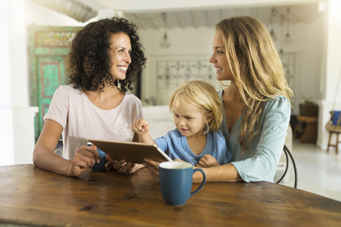 Zwei lächelnde Frauen mit einem Kind, das ein Tablet am Küchentisch benutzt, lizenzfreies Stockfoto