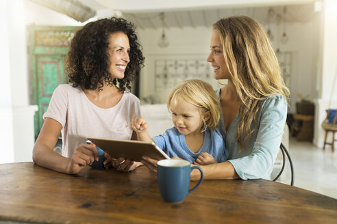 Zwei lächelnde Frauen mit einem Kind, das ein Tablet am Küchentisch benutzt - SBOF01455
