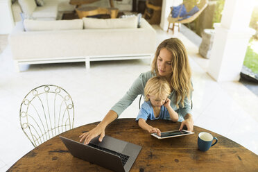 Junge sitzt auf dem Schoß seiner Mutter und schaut auf ein Tablet, während seine Mutter an einem Laptop arbeitet - SBOF01457