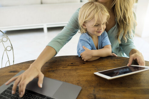 Junge sitzt auf dem Schoß seiner Mutter und schaut auf ein Tablet, während seine Mutter an einem Laptop arbeitet - SBOF01458