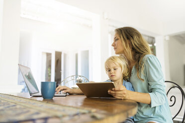 Boy sitting on his mother's lap and looking at a tablet while his mother is working on a laptop - SBOF01459