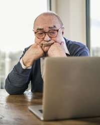 Älterer Mann sitzt am Tisch und schaut auf einen Laptop - UUF12920