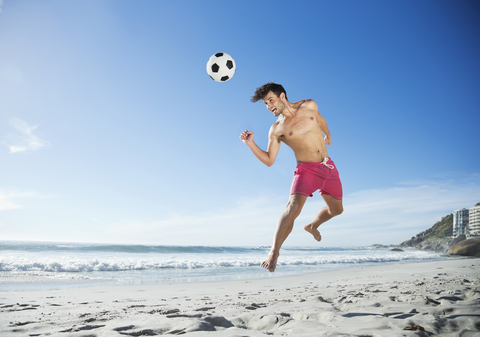 Mann in Badehose köpft Fußball am Strand, lizenzfreies Stockfoto