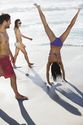 Freunde beobachten Frau im Bikini beim Handstand am Strand - CAIF03538