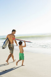 Vater und Sohn tragen Surfbrett und Bodyboard am Strand - CAIF03595