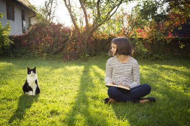 Smiling girl sitting on meadow with a book next to cat - LVF06751