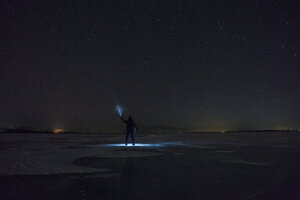 Russia, Amur Oblast, silhouette of man with blue ray standing on frozen Zeya River at night under starry sky - VPIF00381