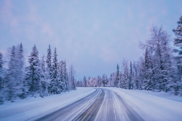 Abgelegene Winterstraße durch schneebedeckte Waldbäume vor blauem Himmel, Lappland, Finnland - CAIF04151