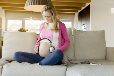 Schwangere Frau sitzt auf der Couch und hält Kopfhörer an ihren Bauch - BMOF00007