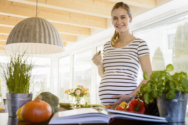 Portrait of smiling pregnant woman in kitchen at home eating cucumber - BMOF00009