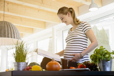 Smiling pregnant woman reading book in kitchen at home - BMOF00026