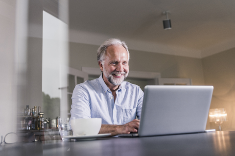 Porträt eines glücklichen reifen Mannes, der zu Hause am Tisch sitzt und einen Laptop benutzt, lizenzfreies Stockfoto