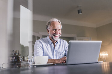 Porträt eines glücklichen reifen Mannes, der zu Hause am Tisch sitzt und einen Laptop benutzt - UUF12922