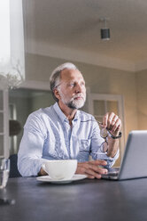 Portrait of pensive mature man sitting at table with laptop looking at distance - UUF12924