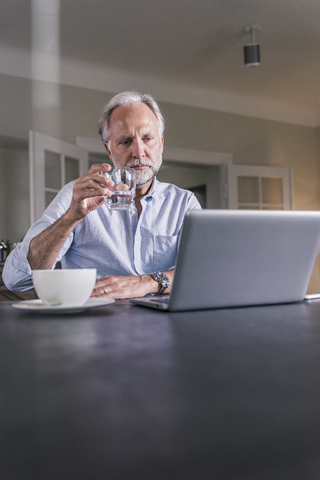 Nachdenklicher reifer Mann sitzt am Tisch mit einem Glas Wasser und schaut auf einen Laptop, lizenzfreies Stockfoto