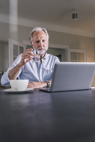 Pensive mature man sitting at table with glass of water looking at laptop stock photo