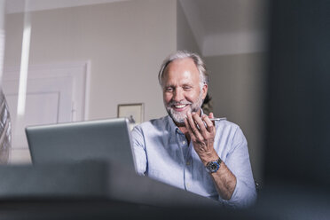 Porträt eines lächelnden reifen Mannes, der zu Hause einen Laptop und ein Mobiltelefon benutzt - UUF12928