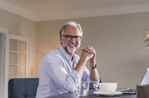 Porträt eines glücklichen reifen Mannes mit Laptop und einer Tasse Kaffee am Tisch in seinem Wohnzimmer, lizenzfreies Stockfoto