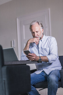 Älterer Mann sitzt am Tisch mit Laptop und schaut auf sein Handy - UUF12933