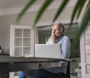 Pensive mature man sitting at table with laptop in his living room - UUF12936