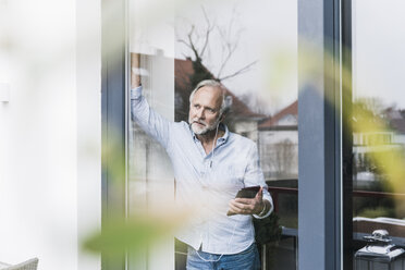 Mature man listening music with earphones and phablet looking out of terrace door - UUF12943