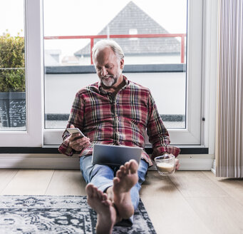 Lächelnder Mann, der sich zu Hause mit Laptop und einer Tasse Milchkaffee auf dem Boden ausruht - UUF12957