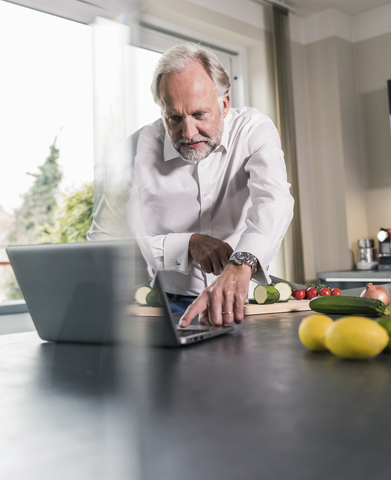 Älterer Mann, der in der Küche Essen zubereitet und dabei einen Laptop benutzt, lizenzfreies Stockfoto