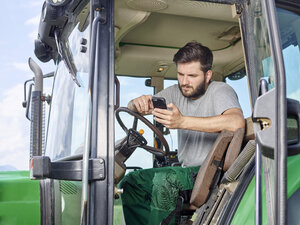 Landwirt sitzt auf einem Traktor und benutzt ein Mobiltelefon - CVF00258