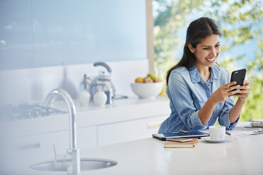 Smiling woman texting with cell phone at kitchen counter - HOXF00089