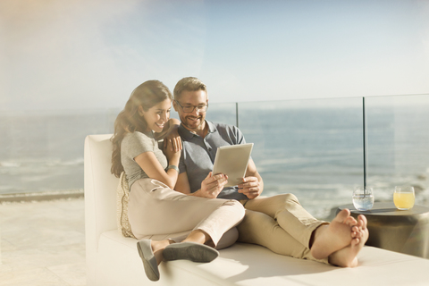 Paar entspannt mit digitalem Tablet auf sonnigem Luxusbalkon mit Meerblick, lizenzfreies Stockfoto