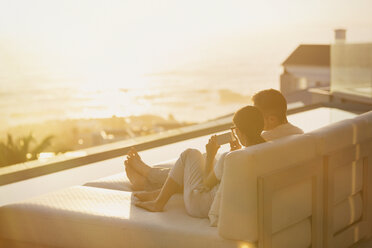 Silhouette couple using cell phone on chaise lounge with sunset ocean view - HOXF00139
