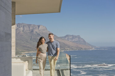 Lächelndes Paar, das auf einem luxuriösen Balkon mit sonnigem Meer- und Bergblick spazieren geht - HOXF00149