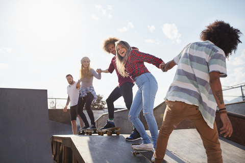 Freunde auf Skateboards halten sich in einer Reihe auf der Rampe im sonnigen Skatepark an den Händen, lizenzfreies Stockfoto