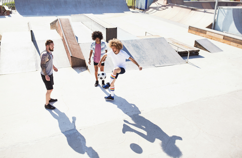 Männliche Freunde spielen Fußball im sonnigen Skatepark, lizenzfreies Stockfoto