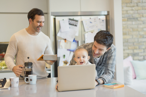 Männliche schwule Eltern und ihr kleiner Sohn benutzen Laptop und Tablet in der Küche, lizenzfreies Stockfoto