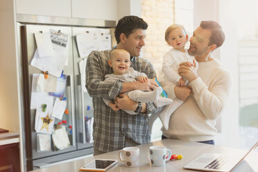 Male gay parents holding baby sons in kitchen - CAIF04302