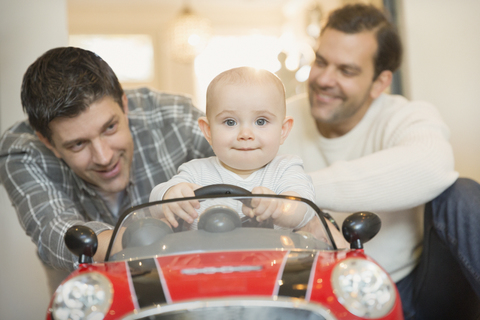 Porträt männlicher schwuler Eltern, die ihren kleinen Sohn im Spielzeugauto schieben, lizenzfreies Stockfoto