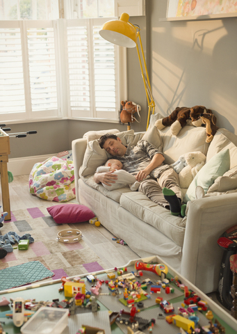 Erschöpfter Vater und kleiner Sohn schlafen auf dem Sofa im unordentlichen Wohnzimmer mit Spielzeug, lizenzfreies Stockfoto