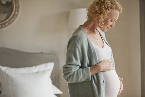 Lächelnde schwangere Frau berührt den Bauch im Schlafzimmer, lizenzfreies Stockfoto