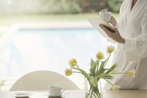 Frau im Bademantel trinkt Kaffee und benutzt ein digitales Tablet am Swimmingpool - HOXF00194