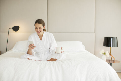 Lächelnde Frau im Bademantel, die auf dem Bett Kaffee trinkt und eine SMS auf ihrem Handy schreibt - HOXF00210