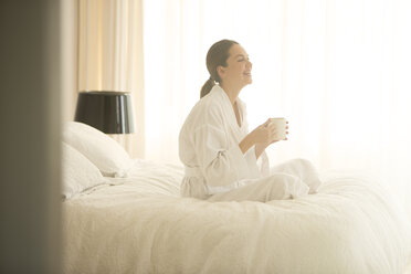 Smiling woman in bathrobe drinking coffee cross-legged on bed - HOXF00219