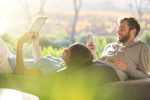 Ein Paar liegt auf einer sonnigen Terrasse und benutzt ein digitales Tablet und ein Mobiltelefon - HOXF00274