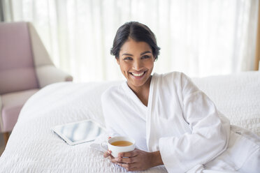 Portrait smiling woman in bathrobe drinking tea on bed - HOXF00288