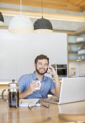 Smiling man drinking coffee and talking on cell phone at laptop - HOXF00309