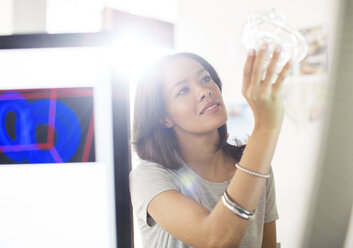 Curious businesswoman examining glass in sunny office - HOXF00337