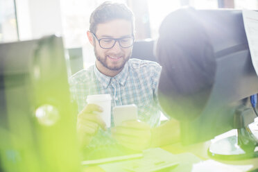 Kreativer Geschäftsmann, der Kaffee trinkt und mit seinem Handy am Computer SMS schreibt - HOXF00351